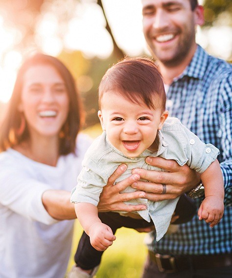 Smiling mother and father holding laughing baby outdoors after preventive dentistry in Austin